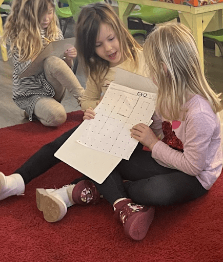 Two girls sit on carpet and look at math grid at micro school.
